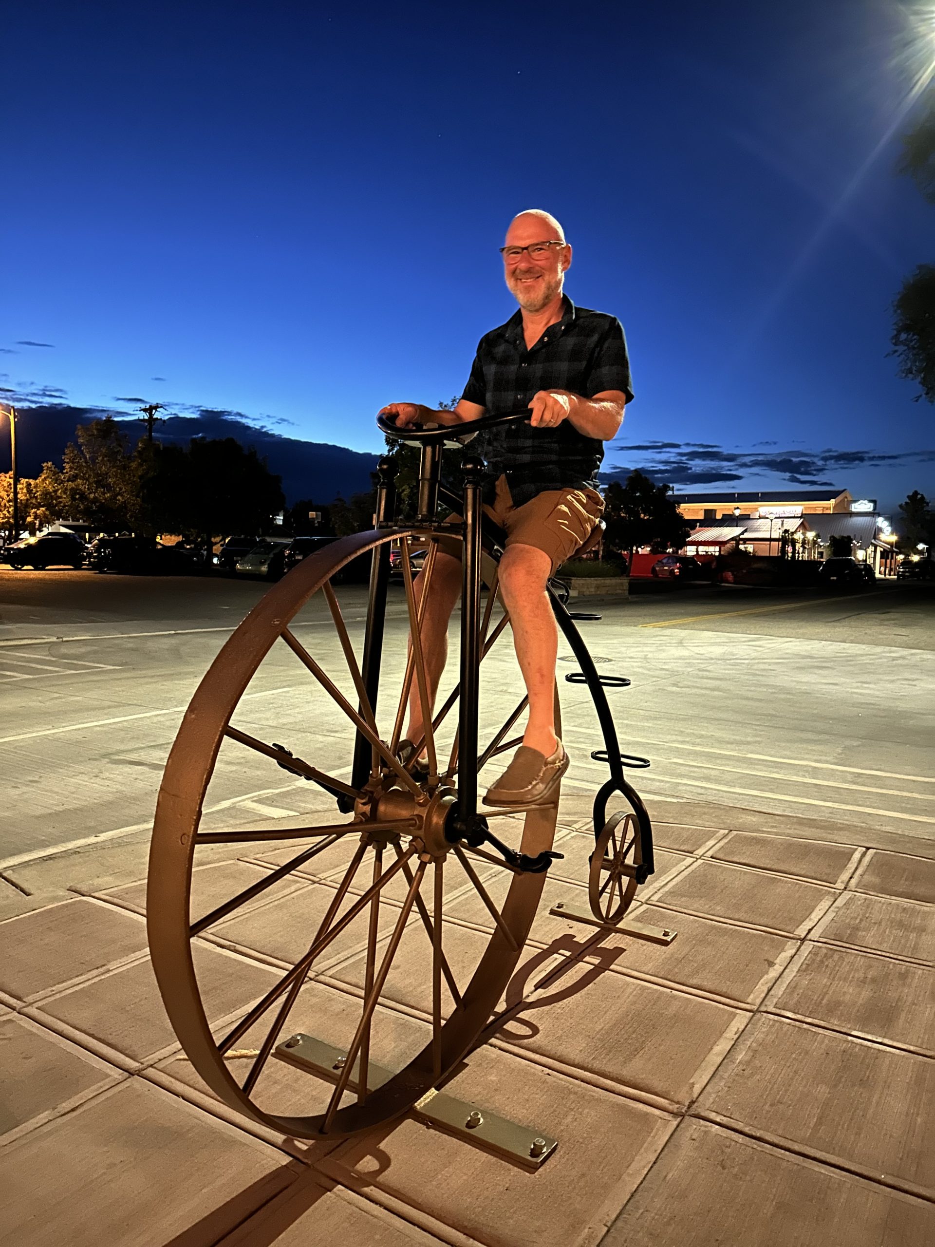 A man sits on a statue of a big wheeled bike at dusk.